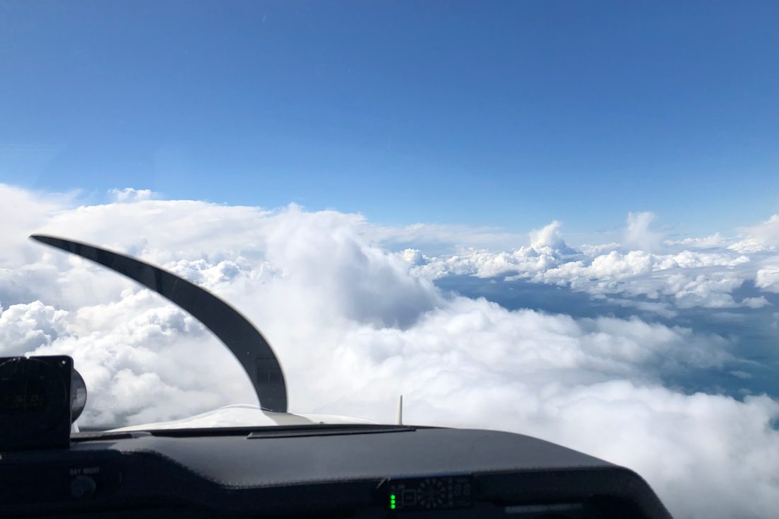 View from the cockpit of a light aircraft above towering cumulus cloud. A propeller blade curves across the left of the frame against a clear blue sky.